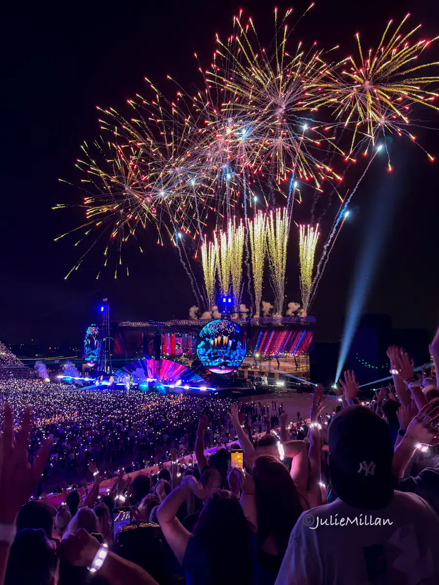 Fireworks explode in colorful bursts above Rogers Stadium during a Coldplay show in Toronto, lighting up the night sky. The stage glows with bright visuals and thousands of fans in the stands cheer with their arms raised, many wearing glowing wristbands. The whole scene feels festive and magical.