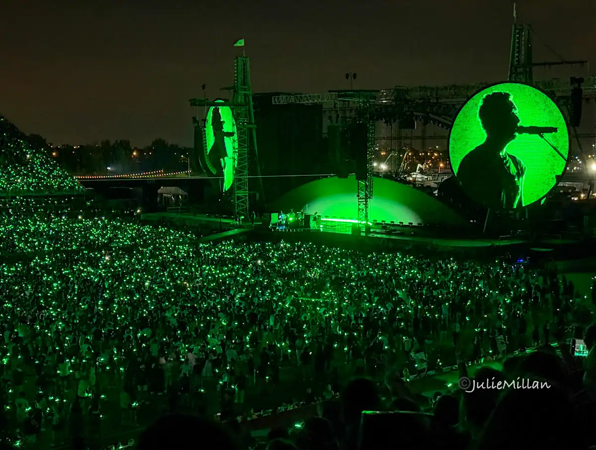 A sea of green lights from fans’ wristbands glows across a packed stadium during a Coldplay concert in Toronto. A silhouette of the lead singer singing into the microphone is projected on a giant circular screen, creating a dramatic, immersive scene. The stage lighting bathes the whole arena in vibrant green.