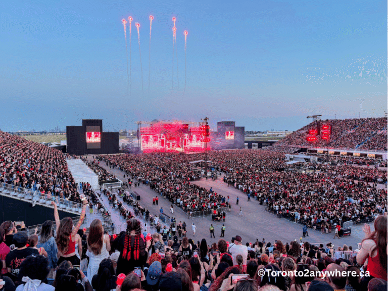 A packed Rogers Stadium at Downsview in Toronto during dusk, with thousands of fans cheering and holding up light sticks as red fireworks shoot up above a brightly lit stage. The energy is electric with people filling both the floor and grandstand seats, set against a clear evening sky.