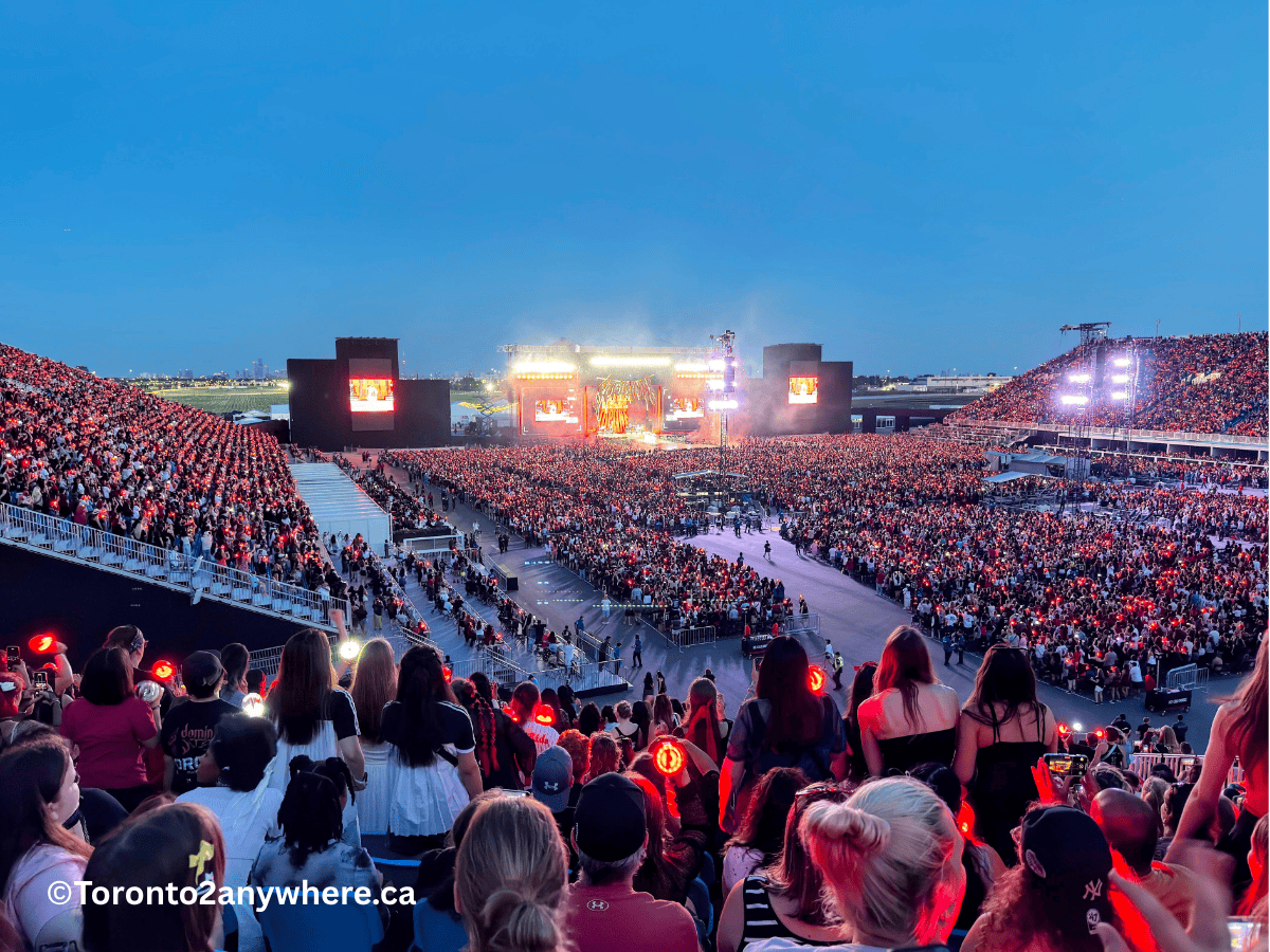 A massive crowd fills Toronto's new Rogers Stadium at Downsview Park during dusk, with fans waving light sticks and watching a brightly lit stage show. The stadium glows under blue twilight, creating an electric atmosphere with packed grandstands and an excited audience.