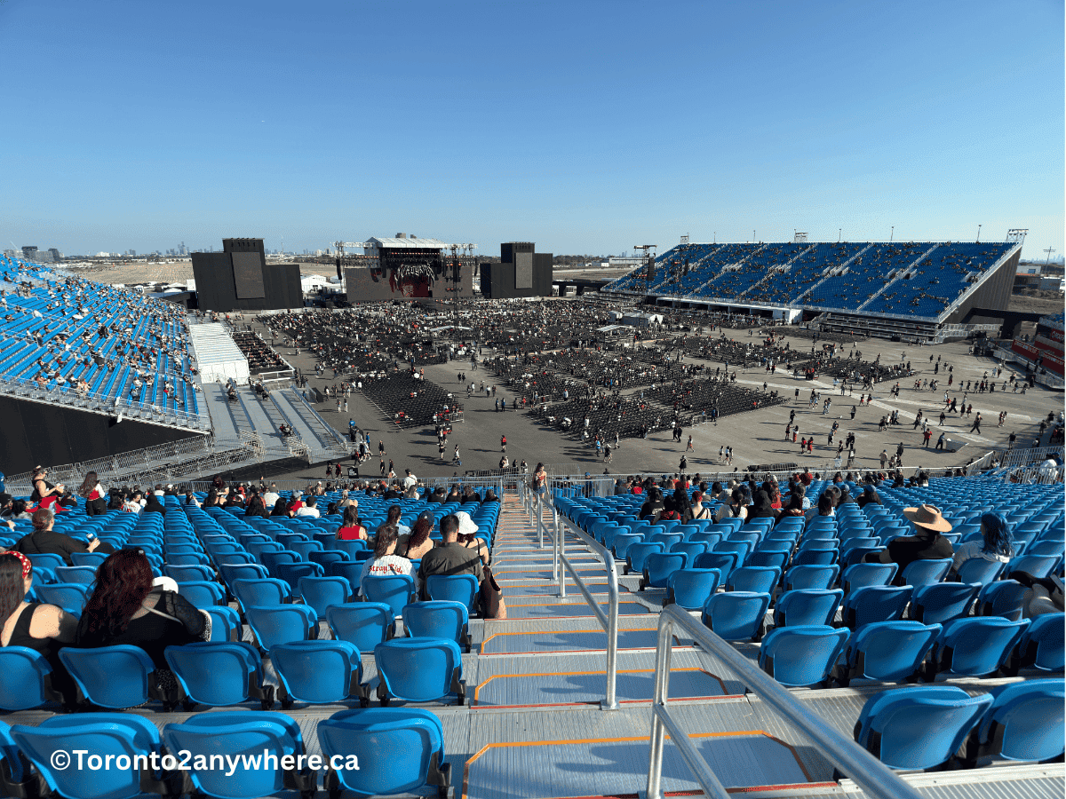 A wide daytime at the new Rogers Stadium showing the stage area, sparsely filled floor seats, and expansive blue grandstands starting to fill up before a concert. People are gradually entering, and the bright sky highlights the scale of the venue.