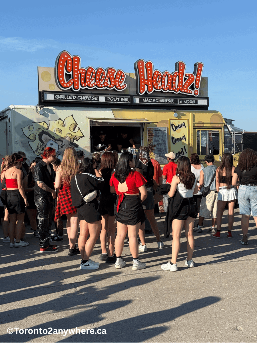 A large crowd gathers in front of a food truck called "Cheese Headz!" at a concert in Toronto, serving grilled cheese, poutine, mac and cheese, and more. People dressed in summer festival outfits wait in line under a clear blue sky, with the sign reading “Ooey Gooey Good!”