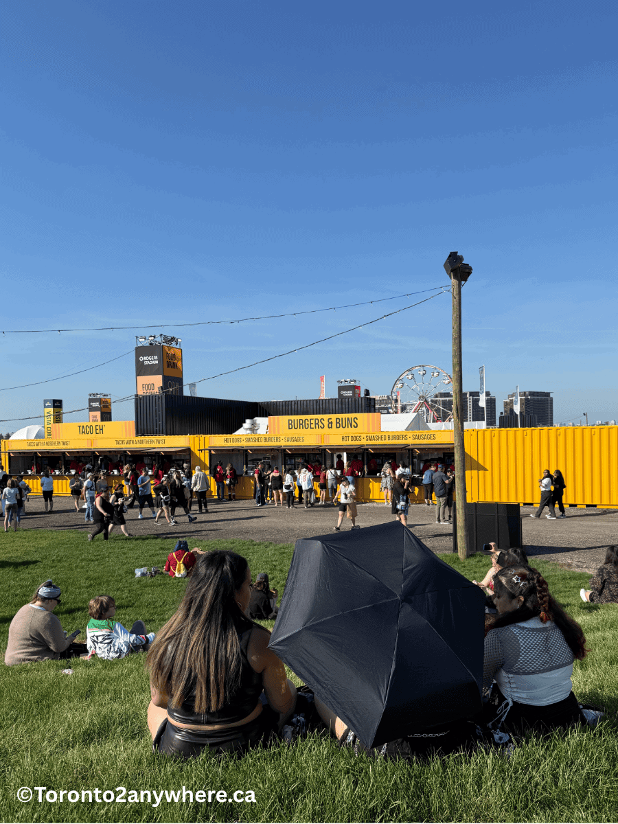 Concert fans relax on a grassy lawn near bright yellow food vendor stalls labeled “Taco Eh” and “Burgers & Buns,” with a Ferris wheel and city buildings visible in the background under a clear blue sky. Some people sit under an umbrella, enjoying the lively atmosphere ahead of a summer concert at Rogers Stadium in Toronto.