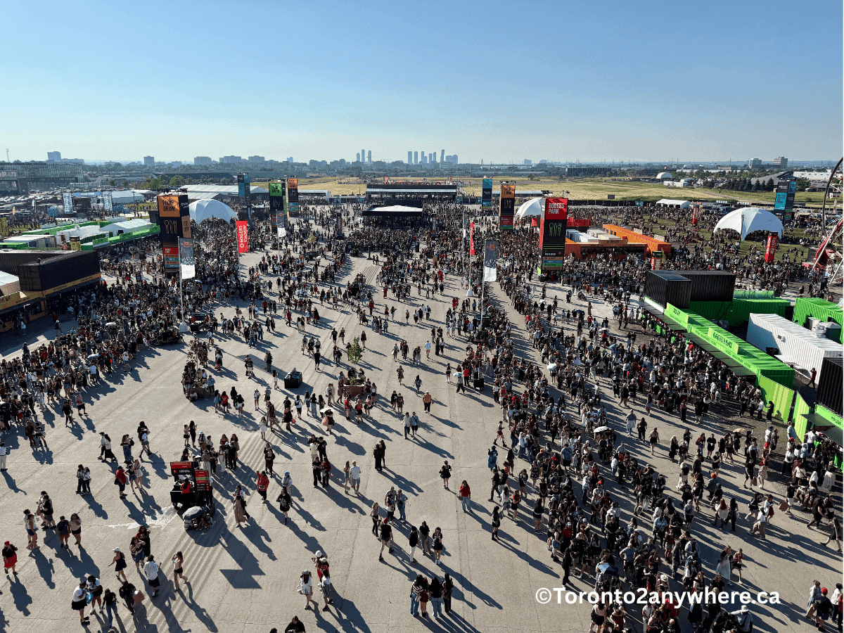 A large aerial view of festival grounds outside Rogers Stadium in Toronto, bustling with people exploring food stalls, merchandise stands, and entertainment areas on a sunny day. Colorful signage and container structures stand out, with the city skyline in the distance.