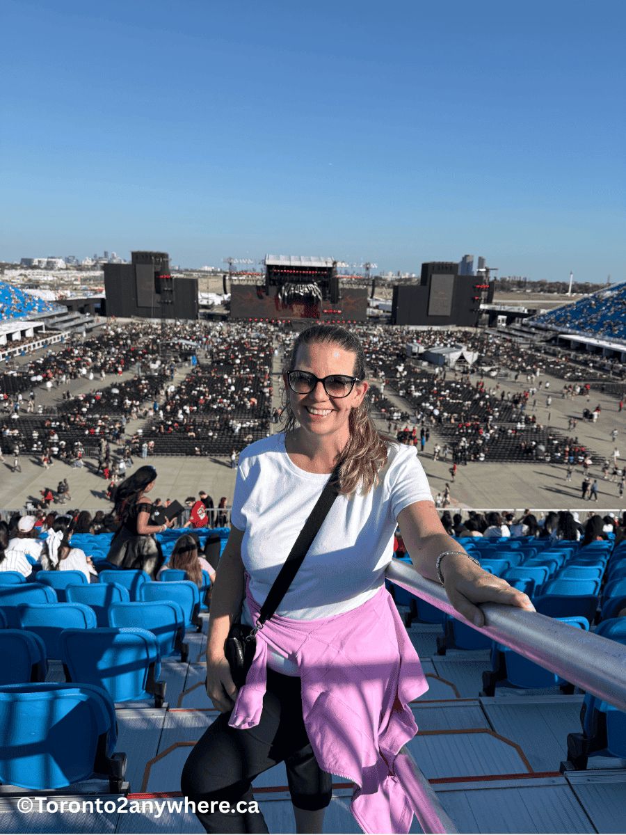 A smiling woman poses in front of bright blue seats inside the Rogers concert venue at Downsview Park in Toronto with the stage and floor seating area starting to fill up behind her under a sunny sky. The massive stage setup and big screens are visible, with fans getting ready for the concert.
