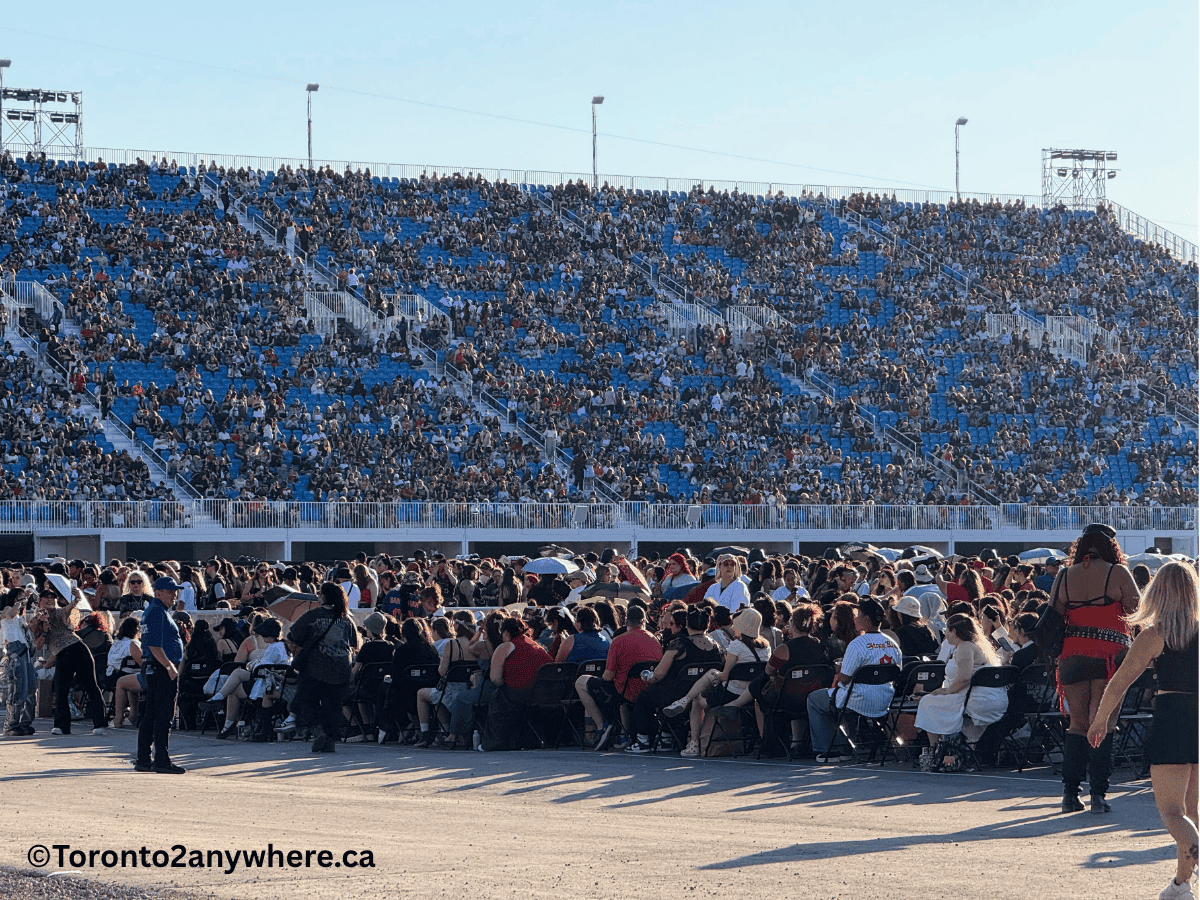 A close-up view of fans sitting and standing in floor seats and packed blue grandstands at Toronto's Rogers Stadium during daylight. The scene is lively with people chatting and moving around, while security staff watch nearby.