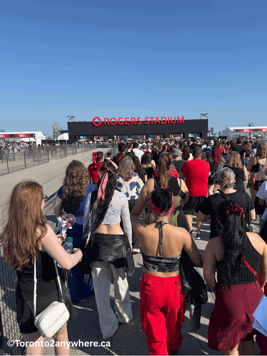 A large crowd of people lines up to enter Rogers Stadium in Toronto through Gate 1 on a bright, sunny day. The group, dressed in casual and trendy outfits, moves toward the black entrance building marked with bold red “Rogers Stadium” signage.