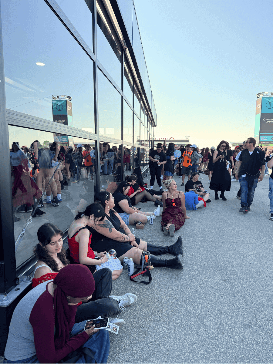 A group of concertgoers sit on the ground along a shaded glass wall outside at Rogers Stadium, resting and checking their phones or chatting. People are dressed in casual and festival outfits, enjoying a break from the sun on a bright, clear day.