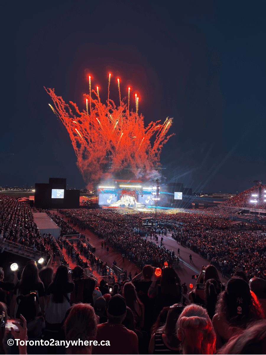 A packed nighttime concert at Rogers Stadium in Toronto, with a massive audience watching a brightly lit stage while red fireworks explode dramatically overhead. Fans hold up phones and light sticks, capturing the electrifying performance.