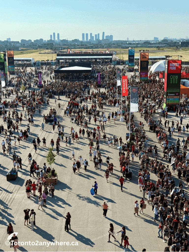 An elevated view of the bustling outdoor entrance area to Rogers Stadium in Toronto, filled with crowds moving between food stalls, signposts, and large event banners. The city skyline and open fields are visible in the background under clear blue skies.
