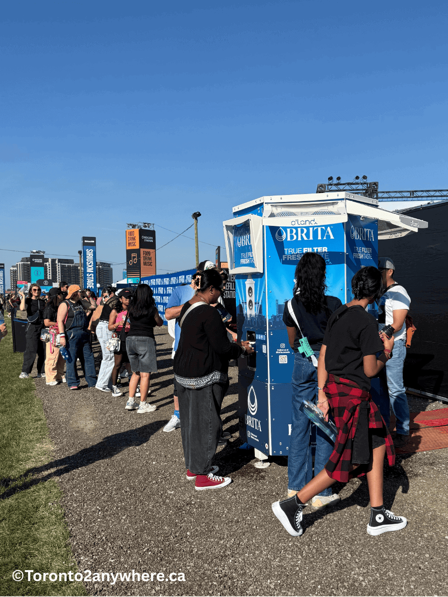 A long line of people waits at a blue Brita water refill station during a sunny concert day at Rogers Stadium, Toronto. Attendees hold reusable bottles and wear casual summer outfits, with signage and stages visible in the background.