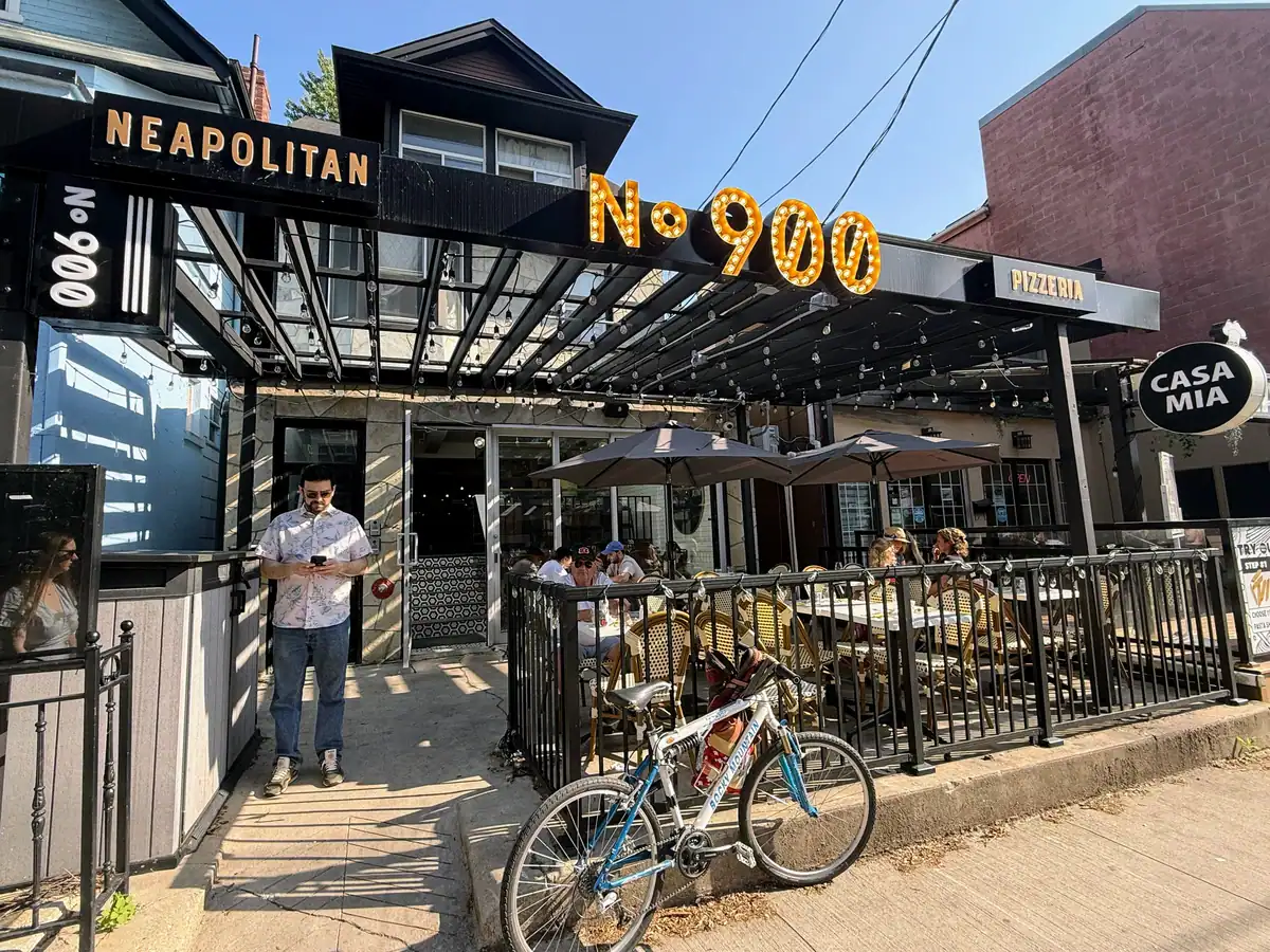 Outdoor view of No. 900 Pizzeria, a Neapolitan-style pizza restaurant with a covered patio area full of diners seated under umbrellas. A man stands by the entrance looking at his phone, and a bicycle is parked against the patio railing in the foreground.