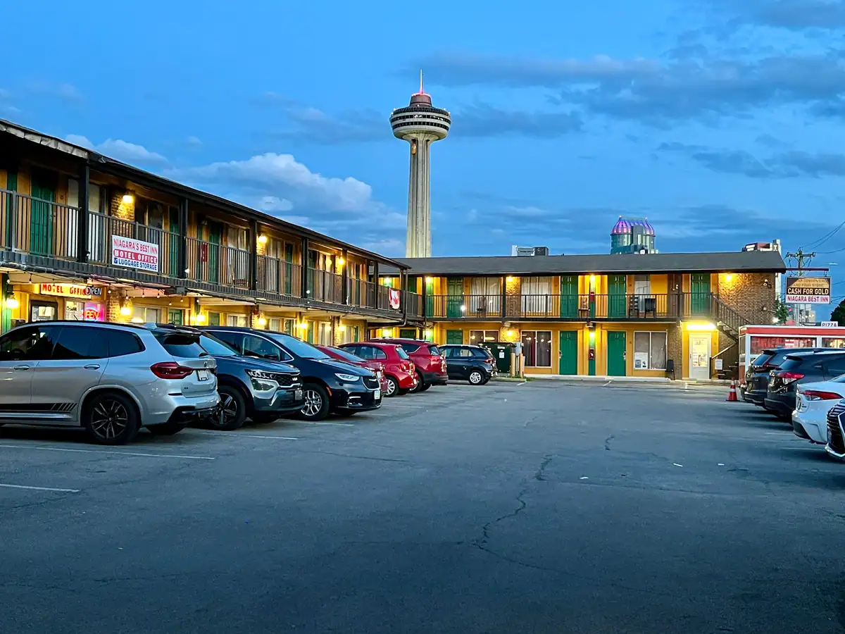 Evening view of the budget Cadillac Motel in Niagara Falls Ontario with cars parked in front and the Skylon Tower lit in the background under a twilight sky.