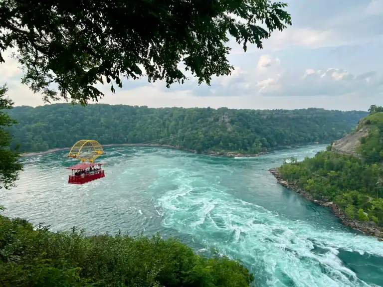 The Whirlpool Aero Car glides over the swirling turquoise rapids of the Niagara River, suspended above the Niagara Whirlpool and framed by lush green forest on both sides. The bright red cable car with a yellow frame contrasts against the dramatic currents and overcast sky.
