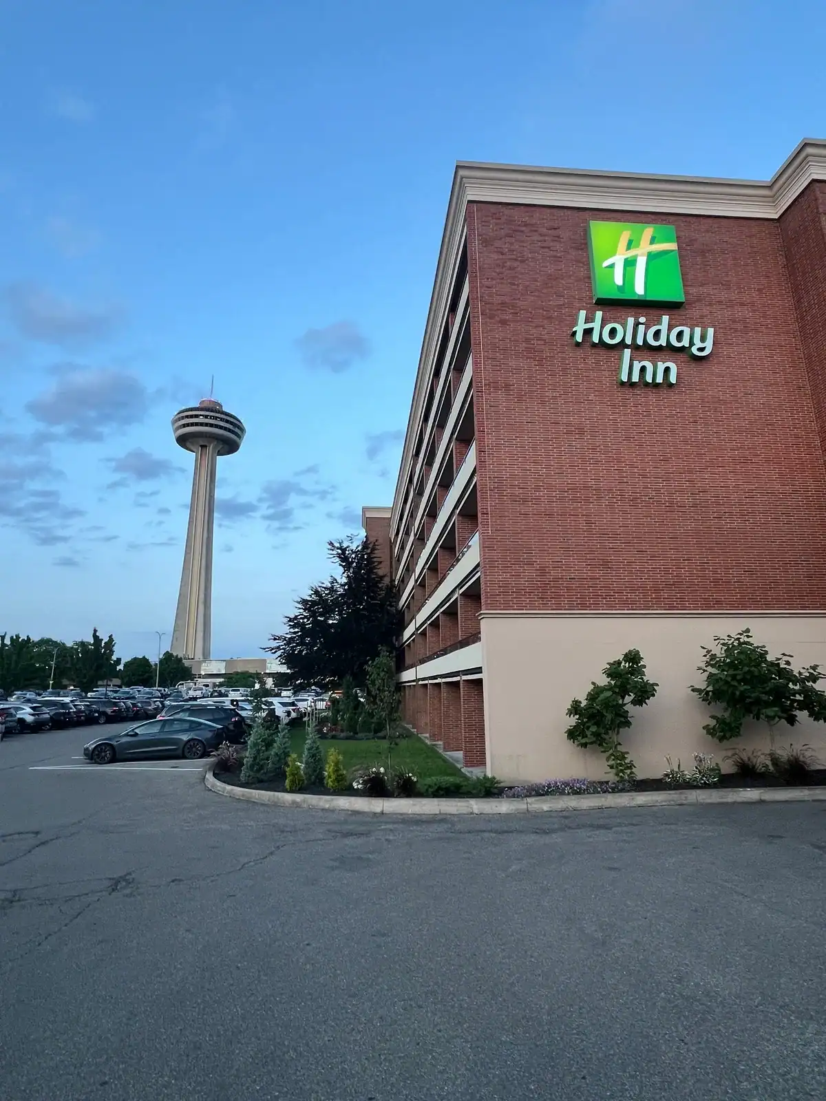 Holiday Inn Niagara Falls with the Skylon Tower visible in the background, a parking lot full of cars, and a well-kept landscaped entrance under a blue sky.