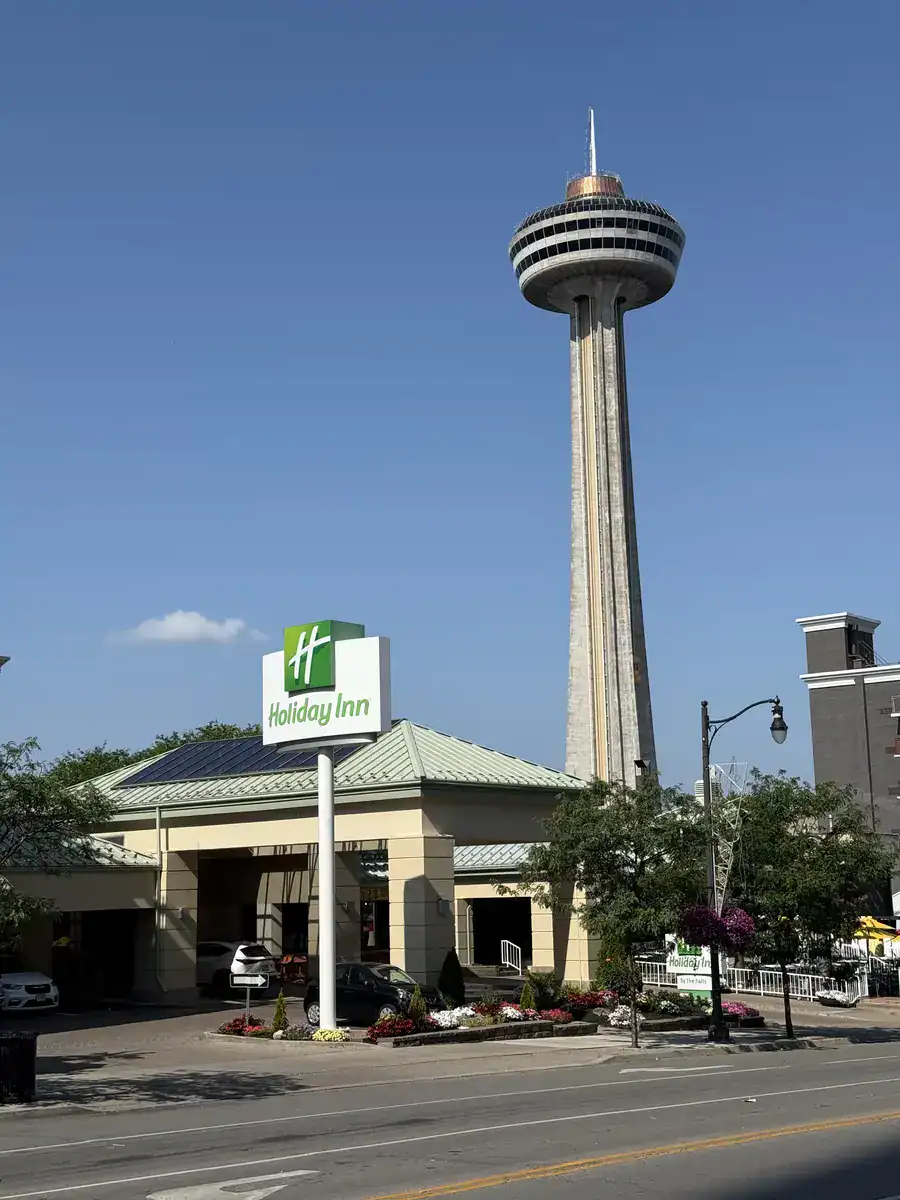 Exterior view of the Holiday Inn on the Falls hotel in Niagara Falls Canada with the Skylon Tower prominently rising behind it against a clear blue sky.