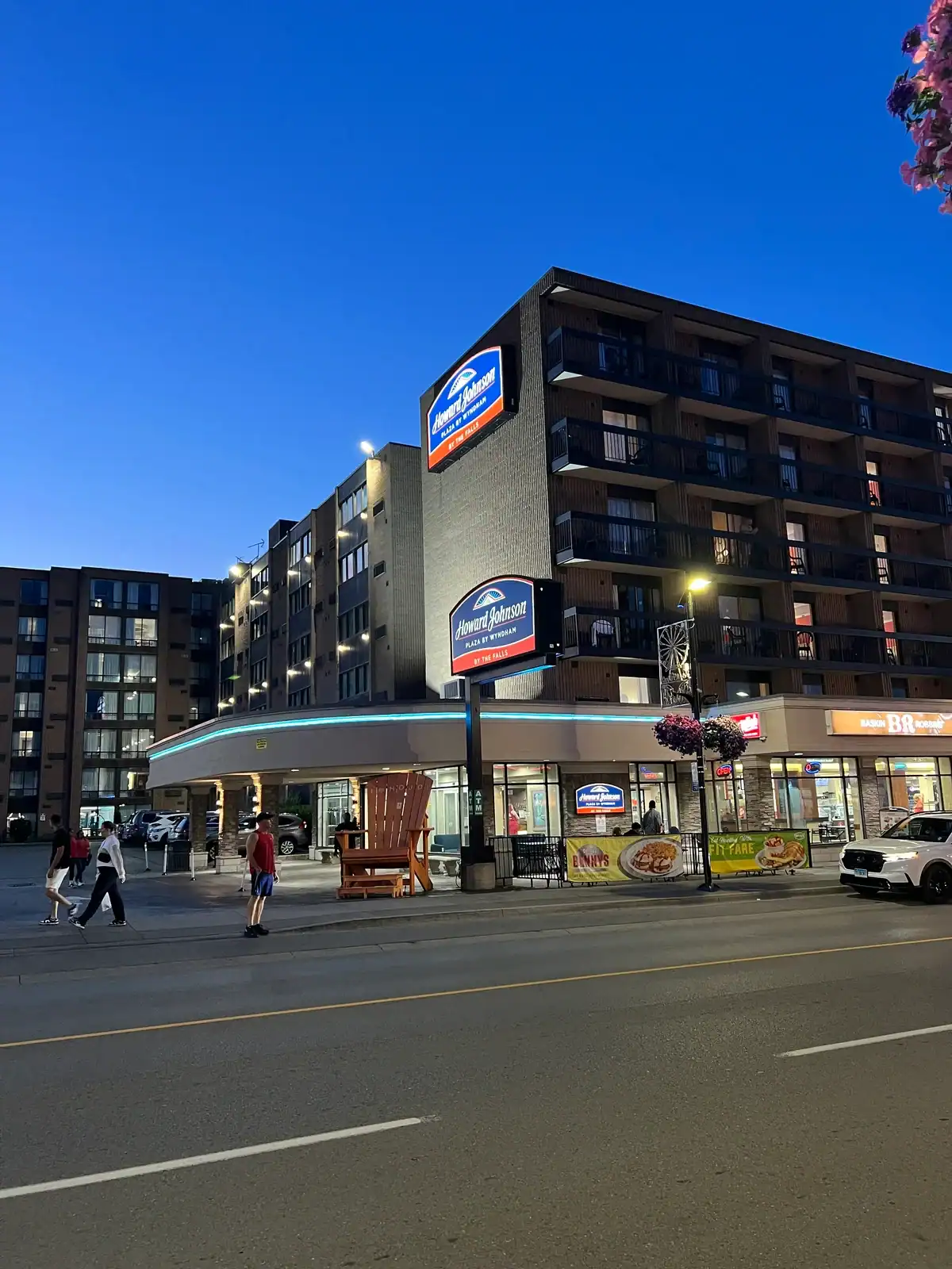 Evening view of the budget Howard Johnson Plaza Hotel by Wyndham in Niagara Falls Ontario Canada with Denny’s restaurant on the ground level and several pedestrians walking by.