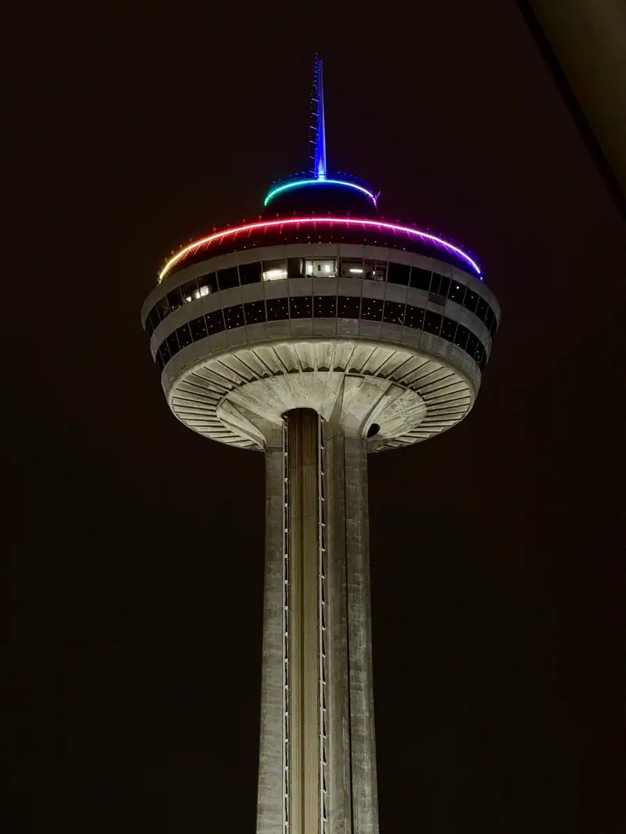 Close-up view of the Skylon Tower in Niagara Falls at night, lit with a rainbow of vibrant LED lights circling the observation deck and spire against a dark sky. The concrete structure and windows are clearly visible under soft illumination.