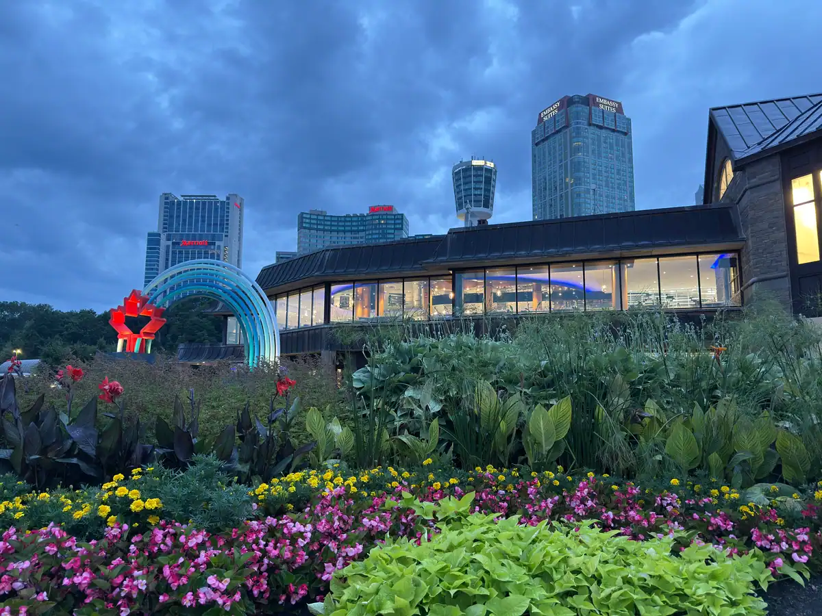 Colourful flower garden in the foreground with a view of the illuminated Niagara Falls Welcome Centre in Canada at dusk, framed by a glowing rainbow arch with a red maple leaf. In the background, fallsview hotels including the Marriott and Embassy Suites tower over the cityscape under dramatic evening clouds.