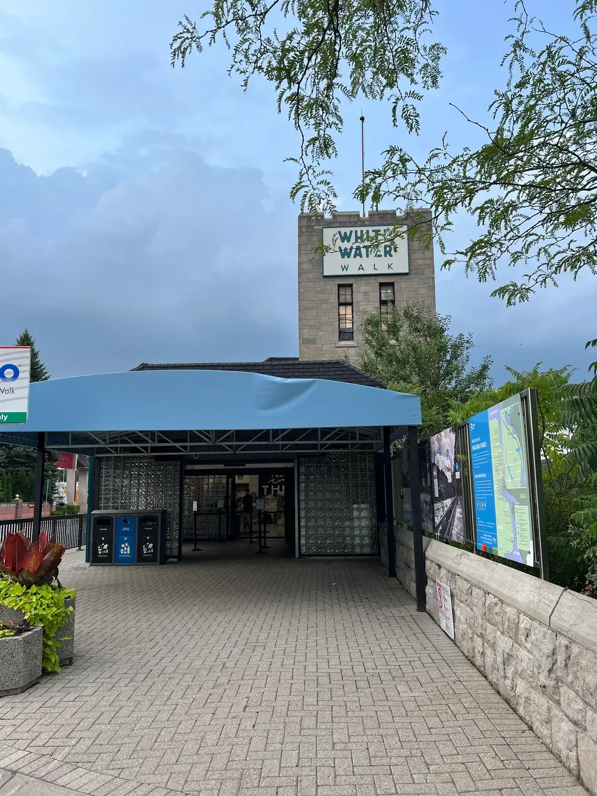 View of the White Water Walk entrance in Niagara Falls, Ontario, with a blue awning and a stone building in the background marked by a sign that reads “White Water Walk”. A tree frames the top of the image under a cloudy sky.