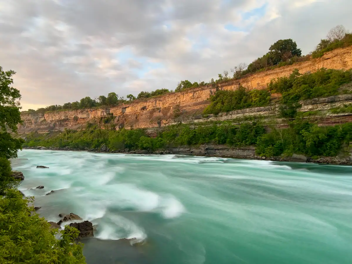 A long exposure captures the Niagara River flowing smoothly past dramatic layered cliffs, with warm light reflecting off the rock face and a cloudy sky above.