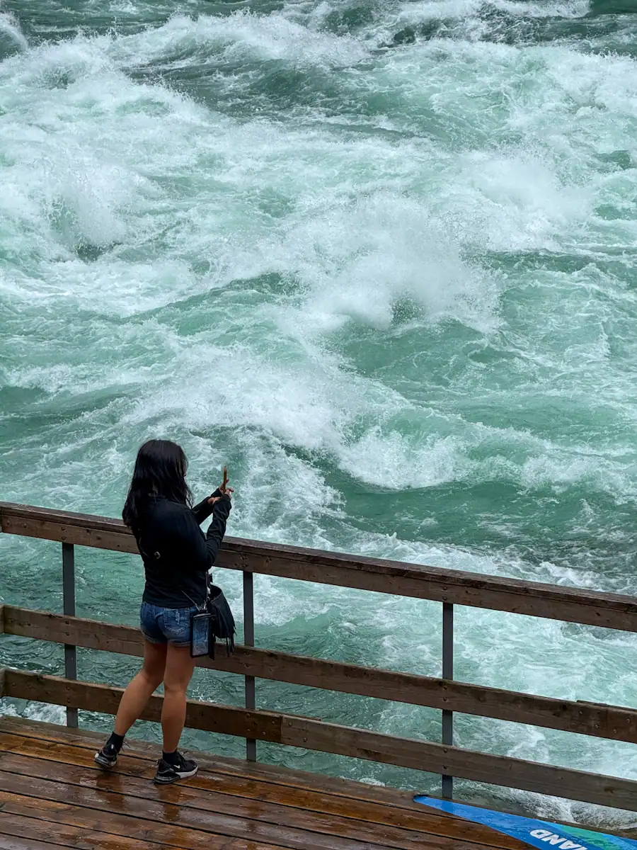 A woman in a black jacket and denim shorts stands on a wooden viewing platform at the White Water Walk in Niagara Falls Canada, taking a photo of the powerful, swirling whitewater rapids below.