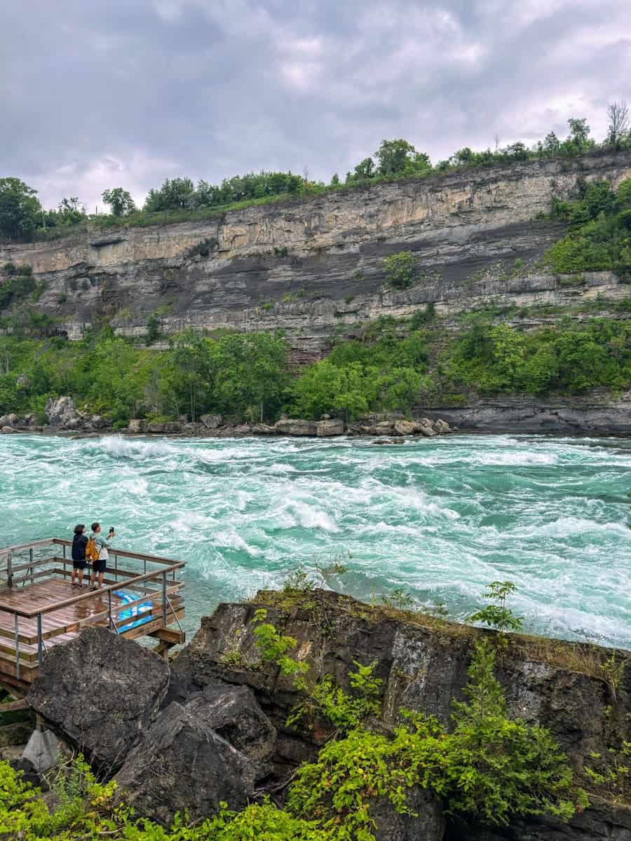 Two visitors stand on a wooden observation platform at the edge of the Niagara River at the White Water Walk, watching the powerful whitewater rapids swirl below under an overcast sky. The rocky cliff face across the river rises steeply, layered with sediment and topped with trees.
