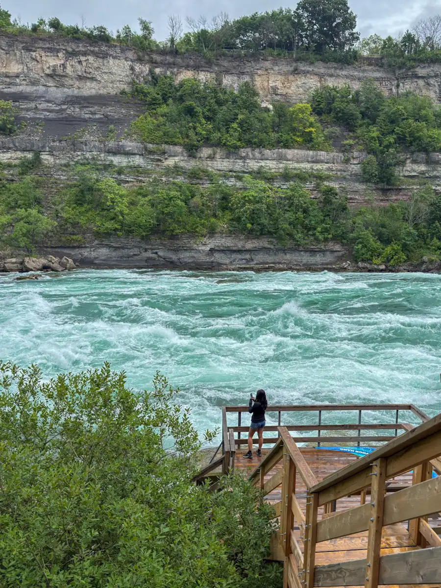 A woman stands on a wooden deck looking out over the turquoise rapids of the Niagara River, with steep rocky cliffs and lush greenery rising on the opposite bank.