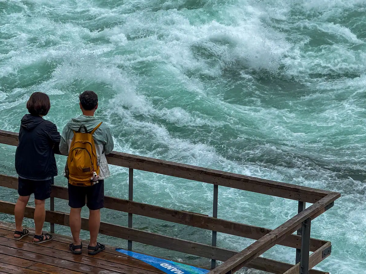 Two visitors wearing jackets and sandals stand at a wooden railing, closely observing the churning whitewater rapids of the Niagara River from the edge of the boardwalk at the White Water Walk.