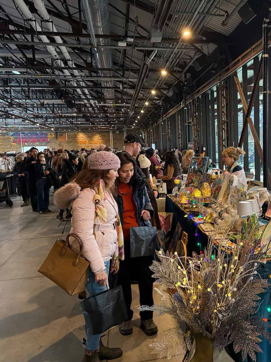 Shoppers browse handmade crafts and holiday decor at a bustling indoor Christmas market at the Evergreen Brickworks in Toronto with twinkling lights and festive displays filling the vendor stalls.