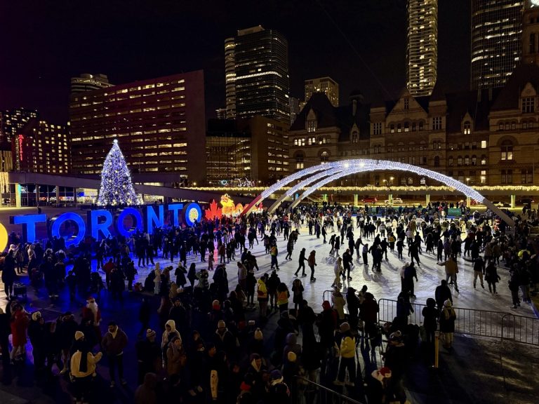 Crowds gather for festive ice skating at Nathan Phillips Square during Christmas in Toronto, with a large illuminated “Toronto” sign, a towering decorated Christmas tree, and glowing holiday arches lighting up the night. Historic and modern buildings frame the bustling winter scene in December.