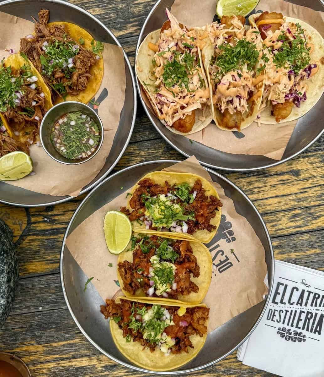 Three plates of vibrant tacos from El Catrin in Toronto’s Distillery District, featuring a mix of slow-cooked meats, fresh cilantro, lime wedges, and colorful slaw on a rustic wooden table.