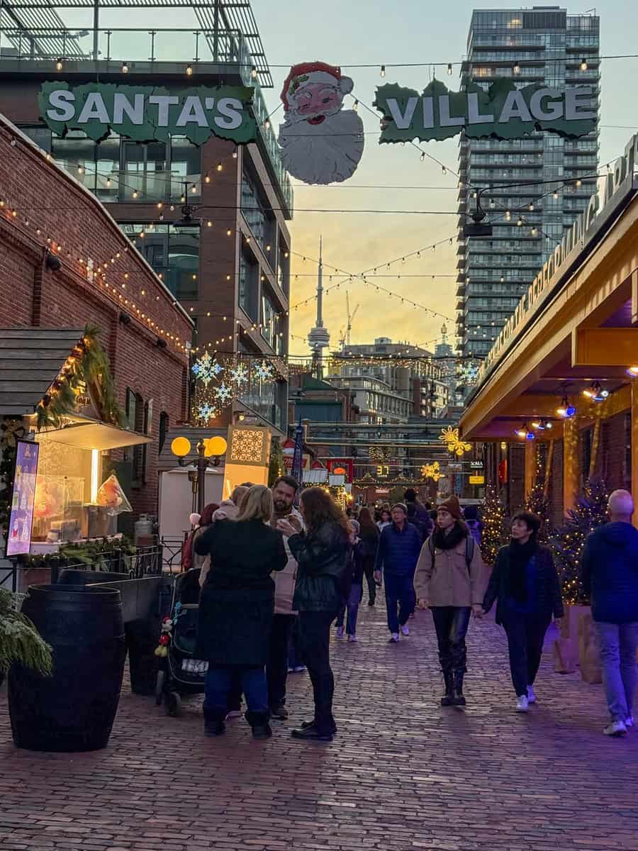 Festive crowd walking through Santa’s Village at the Toronto Distillery District Christmas Market, with twinkling string lights, holiday decorations, and the CN Tower in the distance.