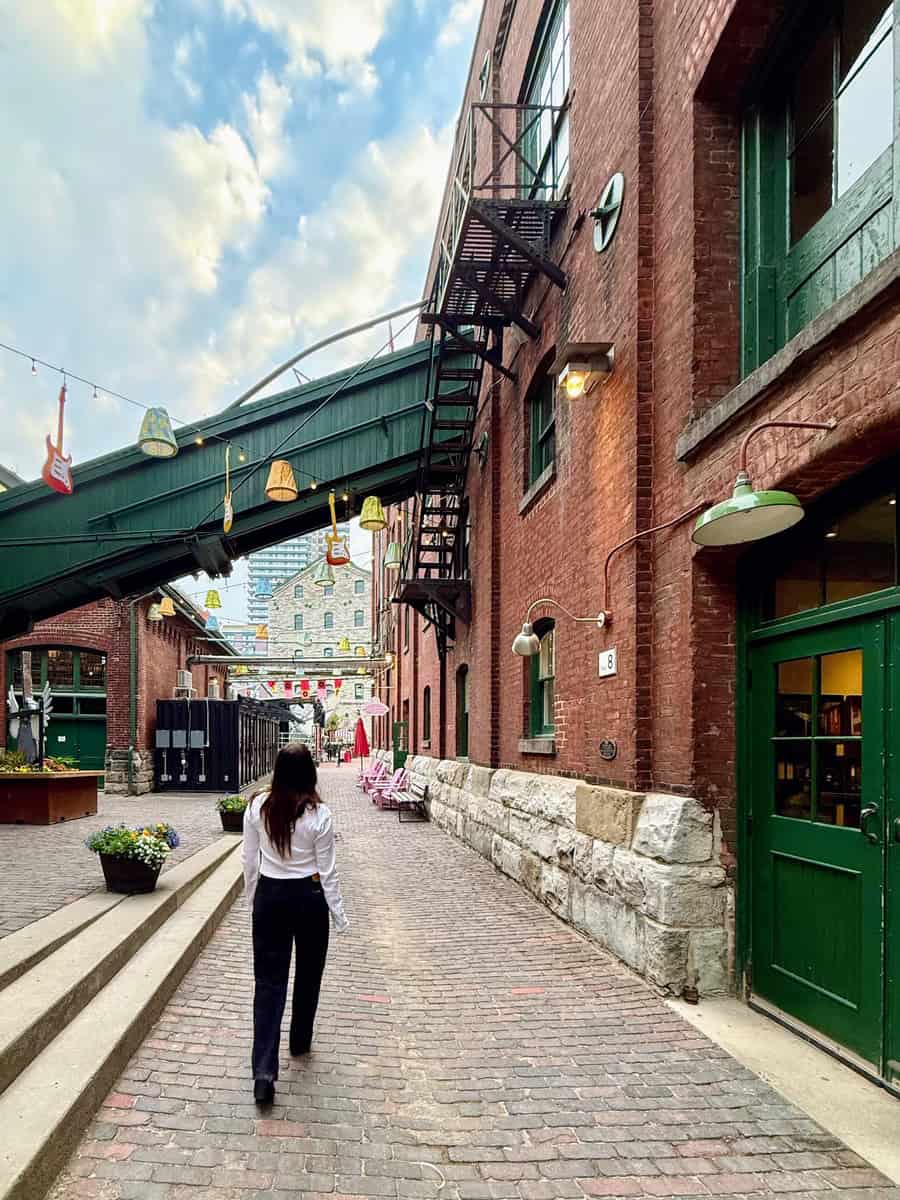 Woman walking along a quiet cobblestone alley in Toronto’s Distillery District, surrounded by historic red brick buildings, green doors, and quirky hanging decor.