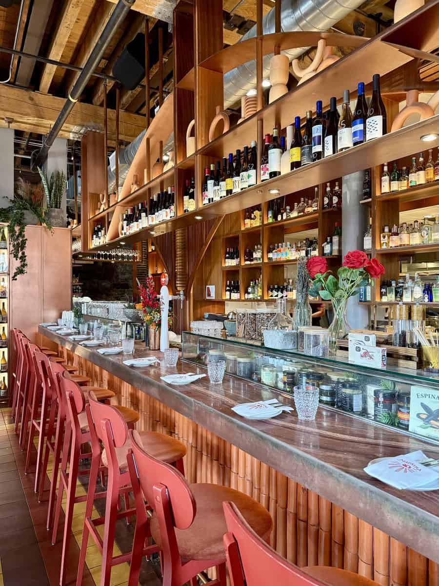 Inside Madrina Tapas Bar , on of the restaurants at the Distillery District, featuring a stylish wooden bar, red bar stools, hanging wine bottles, and a cozy, modern design.