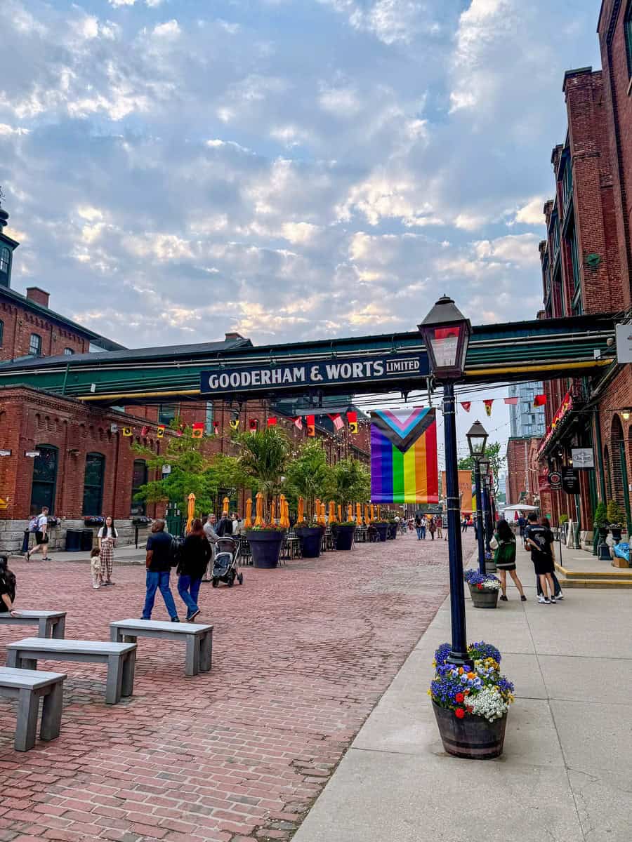 Entrance to the historic Distillery District with Gooderham & Worts signage, rainbow Pride flag, cobblestone streets, and people strolling on a summer day in Toronto.