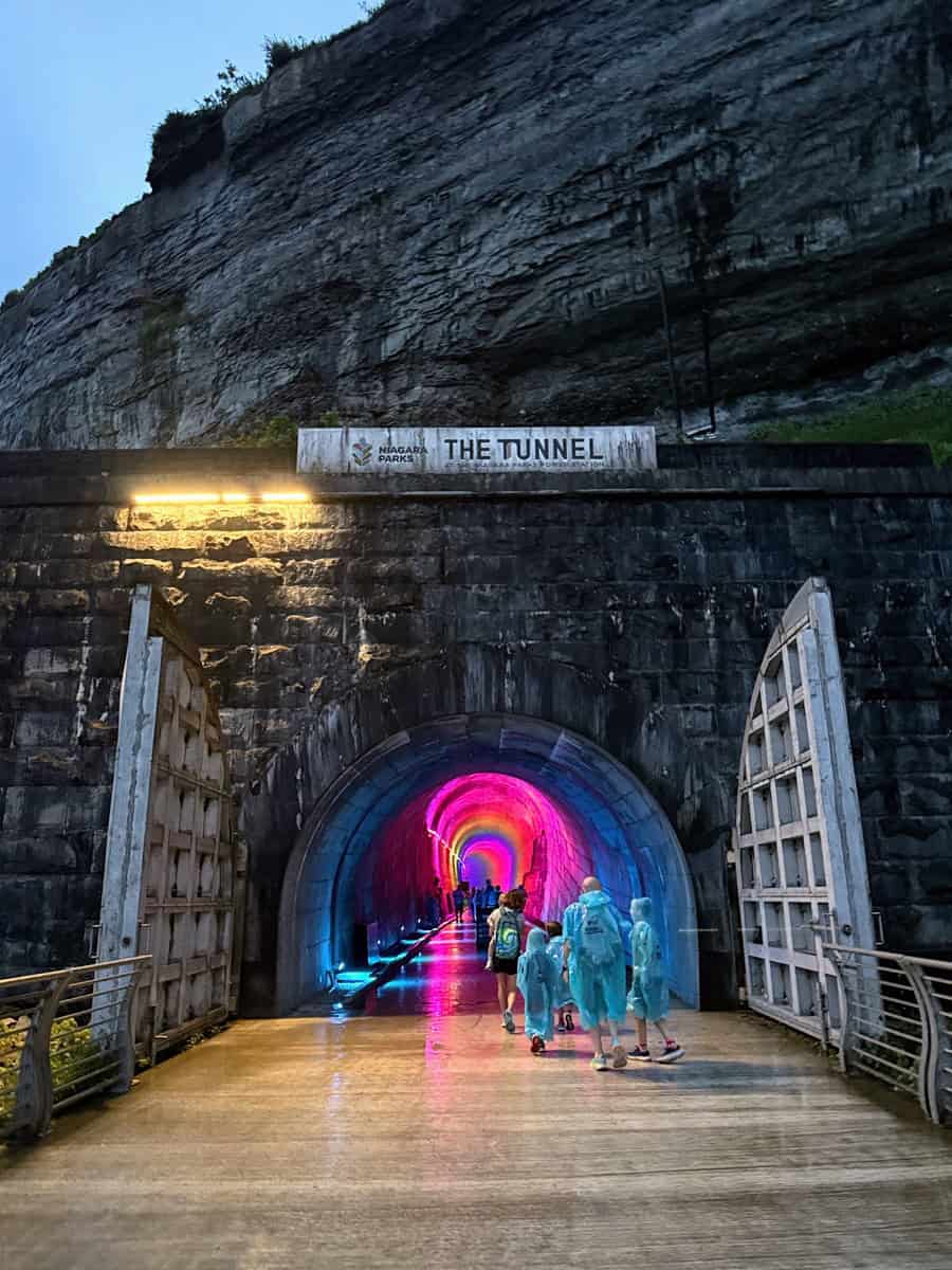 Entrance to the Niagara Parks Power Station Tunnel at dusk, illuminated in pink and blue lights, with visitors in ponchos walking toward the glowing passageway.