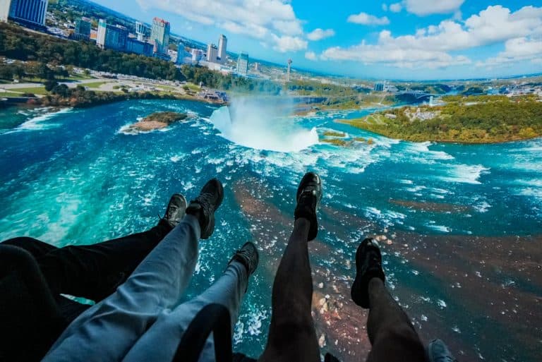 Immersive ride moment inside Niagara Takes Flight, with riders’ legs dangling in front of a sweeping aerial view of Horseshoe Falls and the surrounding city skyline. The cinematic dome screen captures the dramatic mist rising from the falls, making guests feel like they’re soaring high above the water in a thrilling bird’s-eye view. Image credit: Niagara Parks (provided in Media Kit)