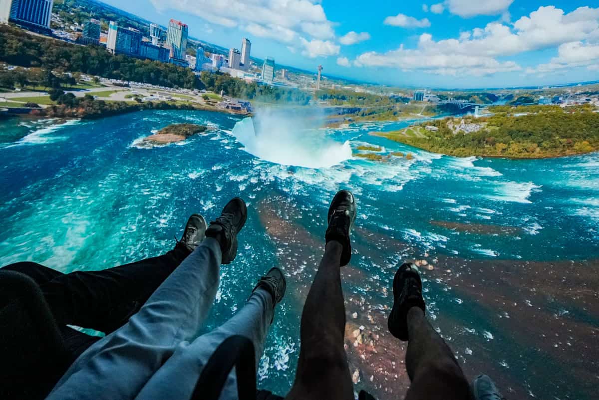 Immersive ride moment inside Niagara Takes Flight, with riders’ legs dangling in front of a sweeping aerial view of Horseshoe Falls and the surrounding city skyline. The cinematic dome screen captures the dramatic mist rising from the falls, making guests feel like they’re soaring high above the water in a thrilling bird’s-eye view. Image credit: Niagara Parks (provided in Media Kit)
