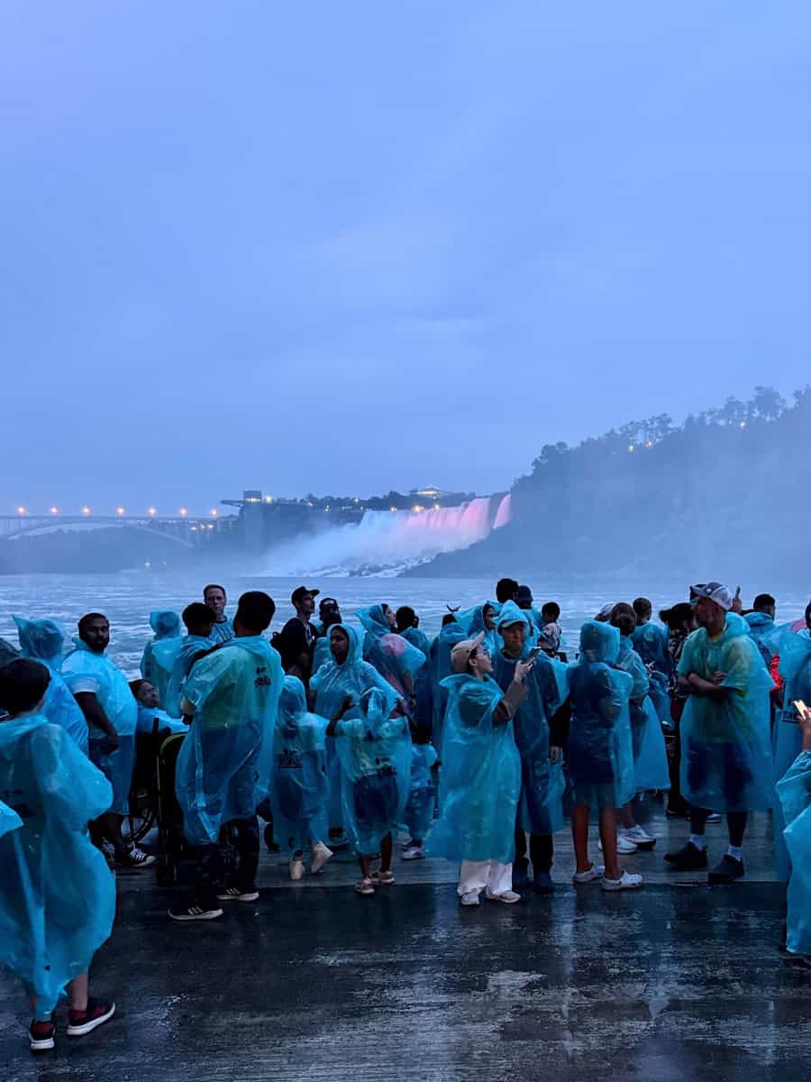 Crowds in blue ponchos gather on the Niagara Parks Power Station observation deck at dusk, watching the illuminated waterfalls and mist rise in the distance.