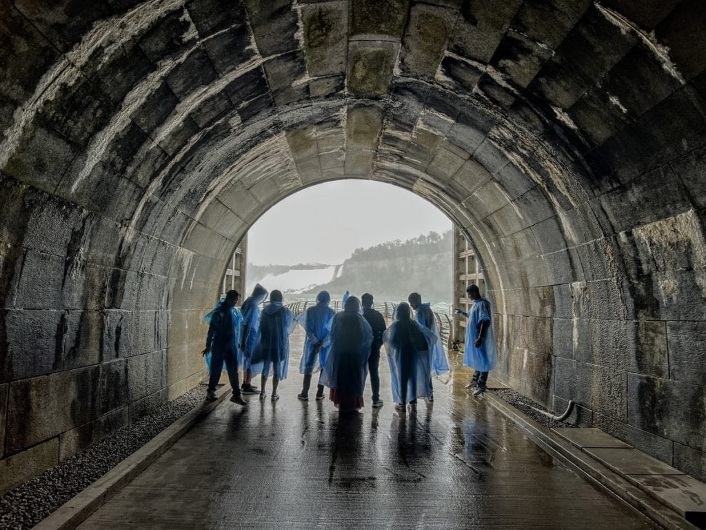 Visitors in blue ponchos stand at the end of the Niagara Parks Power Station tunnel, looking out toward the mist and observation deck by Niagara Falls.