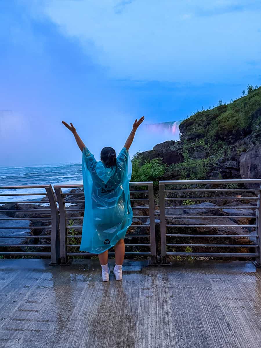 Visitor in a blue poncho raises their arms on the Niagara Parks Power Station observation deck, facing the mist and power of Horseshoe Falls at dusk.