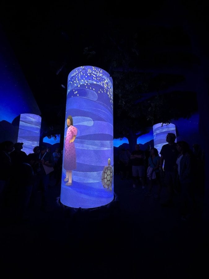 Guests gathered in the dimly lit Ancestors Room, one of the Niagara Takes Flight pre-show experiences. Glowing cylindrical pillars display animated Indigenous artwork — including a woman in a pink robe and a turtle — against a dreamy nighttime forest backdrop. This space tells the Turtle Island creation story, with visuals by Mohawk artist Sara General.
