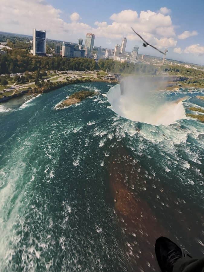 Dramatic in-ride view from Niagara Takes Flight, as riders appear to fly directly over the thundering Horseshoe Falls. The scene captures the sweeping arc of the waterfall with mist rising into the sky, downtown Niagara Falls in the background, and a bird in mid-flight — showcasing just how close and cinematic the experience feels.
