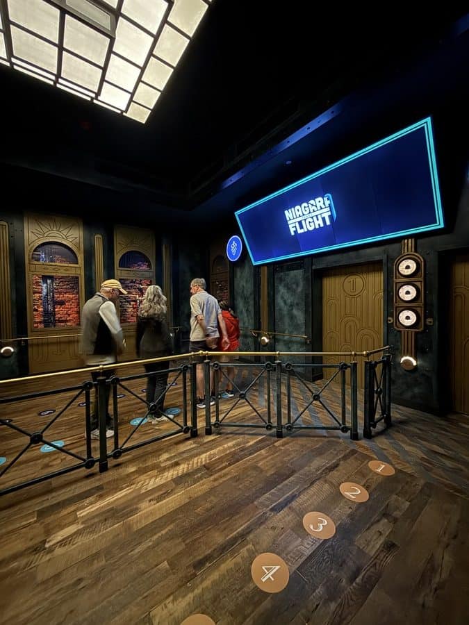 This is the Transporter Room, the final stop before boarding the ride. You can see floor numbers and standing positions, sleek design with dark walls, glowing lighting, and a screen labeled “Niagara Flight.” The design mimics an elevator or futuristic chamber — a detail that ties into the ride’s storytelling of descending underground before launch.