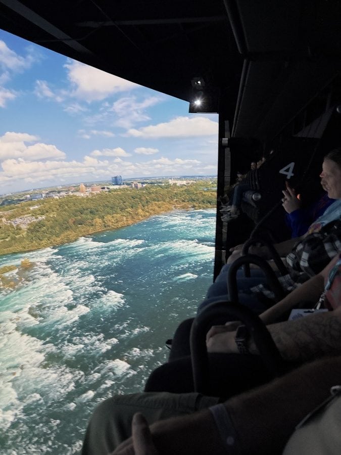 Riders seated in suspended motion chairs inside the Niagara Takes Flight theatre, soaring over the vibrant blue-green waters of the Niagara River. The screen shows a panoramic aerial view of lush green forest, swirling rapids, and distant city skyline — offering a dramatic mid-flight moment during the immersive experience.