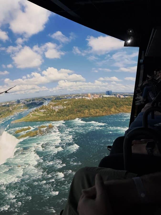 This in-ride photo captures the immersive flying theatre moment when riders soar above Niagara Falls. The screen shows the falls, river, and surrounding landscape, while parts of the seating and viewers’ legs are visible. This is one of the highlights of the experience and gives a strong sense of scale.