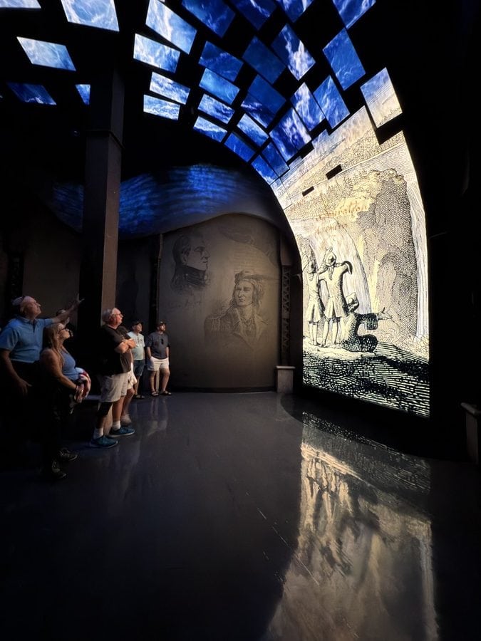 Guests in the Waterfall Room at Niagara Takes Flight standing in front of a dramatic, curved digital screen showing historical sketches of Niagara Falls and explorers. A ceiling of illuminated blue panels mimics flowing water above, creating a multi-sensory environment. This room blends storytelling, technology, and Niagara’s history in a captivating pre-show segment.