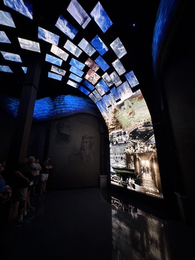 Visitors standing in the Waterfall Room of Niagara Takes Flight, surrounded by a curved wall of black-and-white historical visuals and a digital ceiling filled with vibrant, moving water panels. This pre-show space blends historic imagery with immersive lighting to set the tone for the ride ahead, showcasing the legacy and industrial evolution of Niagara Falls.