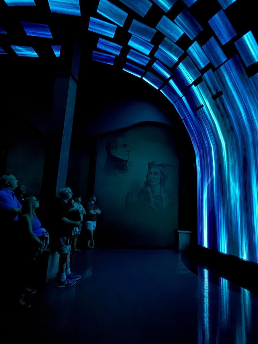 Visitors standing in Niagara Takes Flight’s Waterfall Room, surrounded by an immersive blue-lit ceiling of LED screens and historical imagery projected on arched walls.